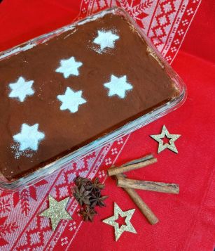 Eine rechteckige Schale mit Tiramisu, dessen Oberfläche mit Kakaopulver bestäubt ist. Darauf sind sechs weisse Sternmuster zu sehen. Die Schale steht auf einem roten Tischtuch mit weissem, weihnachtlichem Muster. Daneben liegen Zimtstangen, Sternanis und goldene Deko-Sterne.  