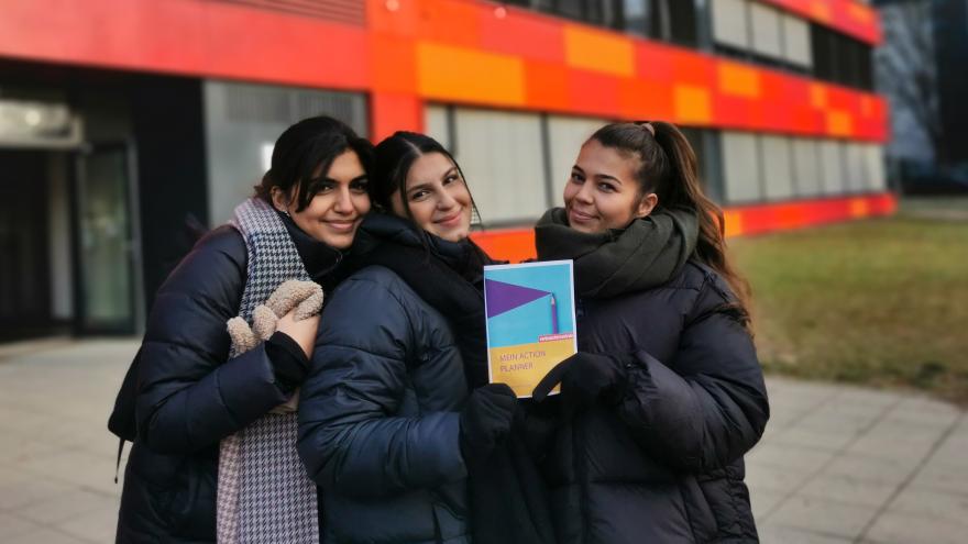 Drei junge Frauen stehen vor einem Gebäude, halten den Action Planner der Verbraucherchecker in den Händen und lächeln.